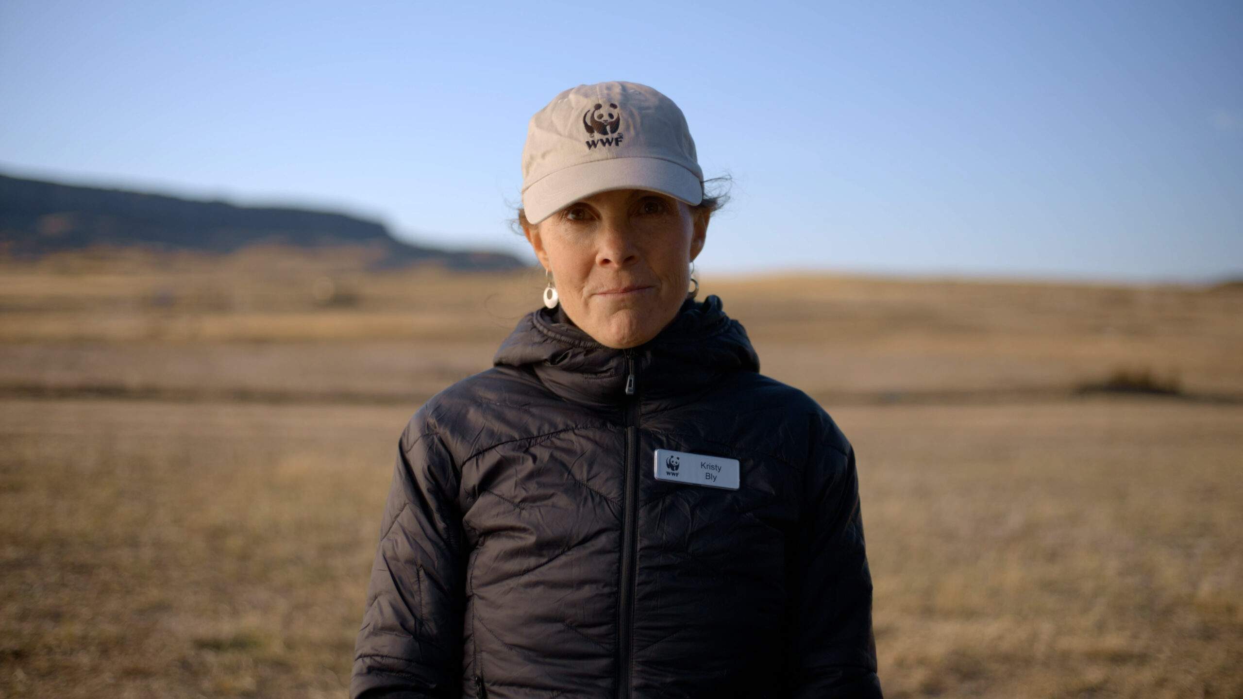 A woman wearing a beige baseball cap and a black jacket stands in a grassy field.
