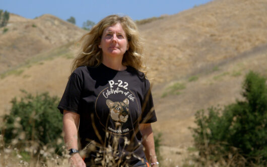 A woman with long, curly blonde hair stands in a black t-shirt that says "P-22."