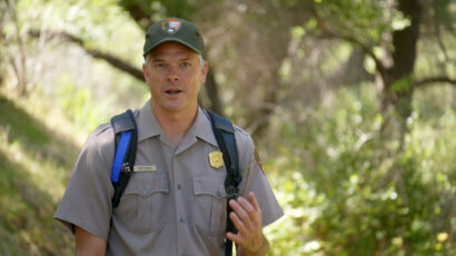 A middle-aged man in a baseball cap and t-shirt stands in front of a forest.