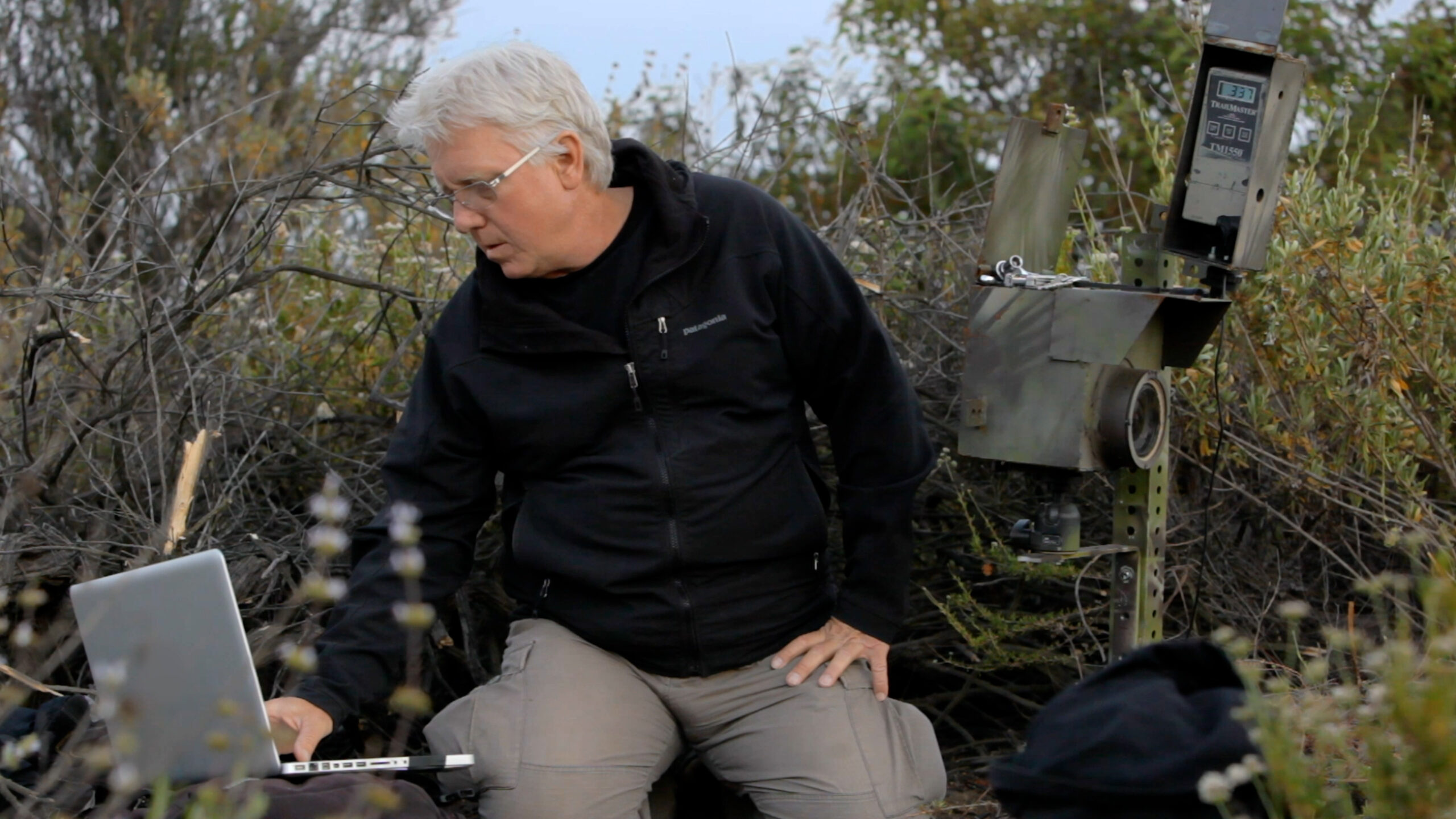 A man with white hair and glasses looks, wearing a black hoodie, sits outside looking at a computer.